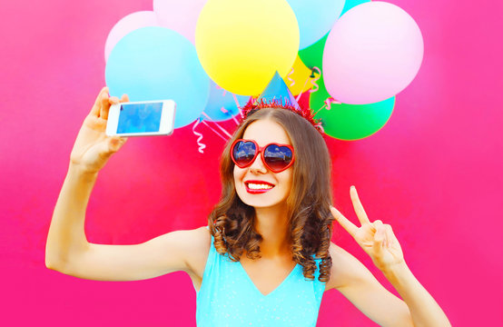 smiling woman in a birthday cap is taking a picture on a smartphone with an air colorful balloons on a pink background