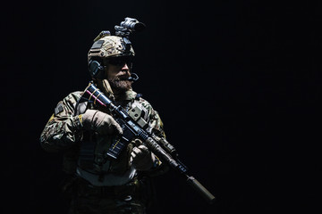 Bearded soldier in Combat Uniforms with weapon, plate carrier and combat helmet are on. Studio shot, dark background