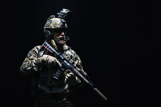 Bearded soldier in Combat Uniforms with weapon, plate carrier and combat helmet are on. Studio shot, dark background