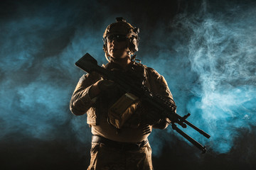 Army soldier in Combat Uniforms with machine gun, plate carrier and combat helmet are on. Studio contour silhouette shot, backlight, dark glowing smoke background