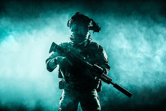 Army soldier in Combat Uniforms with assault rifle, plate carrier and combat helmet are on, Shemagh Kufiya scarf on his neck. Studio contour silhouette shot, backlight, dark glowing smoke background