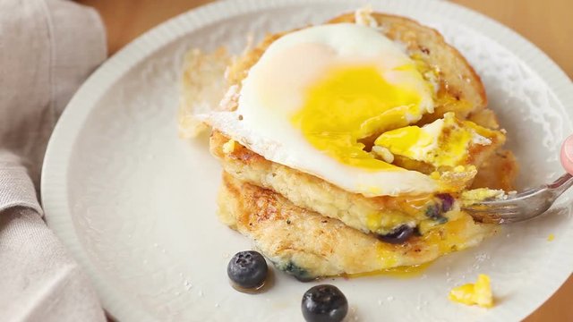 Cutting into a breakfast of blueberry pancakes topped with a fried egg