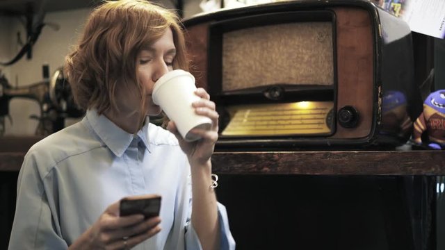 Beautiful young woman is drinking coffee and texting in a pawn shop. Handheld slow motion medium shot