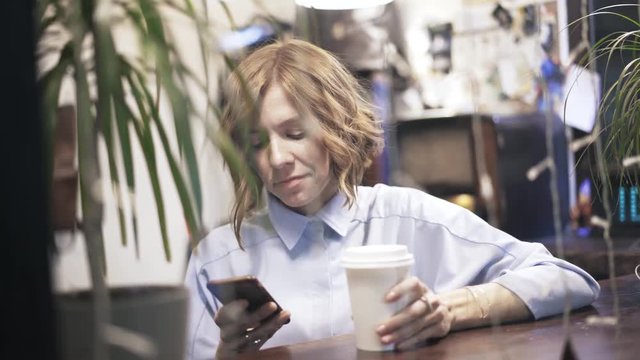 Front view of a beautiful young woman is drinking coffee and texting in a pawn shop. Handheld slow motion medium shot