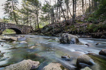 Naklejka na ściany i meble River flows gently along rocks near an arched stone bridge in springtime