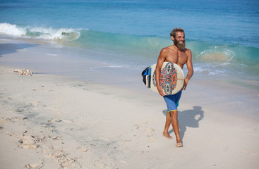 Full-length portrait of an attractive bearded curly man with a surfboard is walking on a beach, located on the right side of the frame
