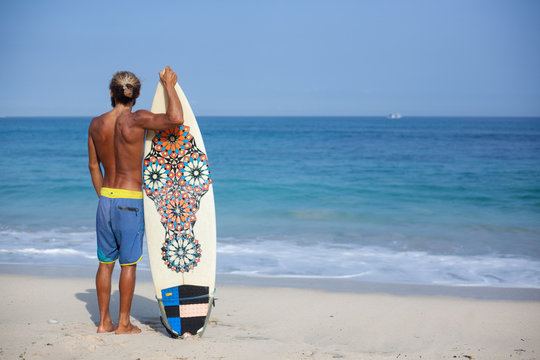 Attractive bearded curly man with a surfboard is standing with his back on a beach and looking to the ocean, located on the left side of the frame