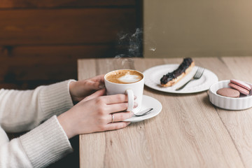 Womans hands wrapped around a cup on wood table with chocolate eclair