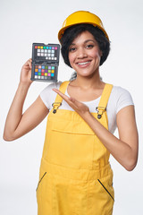 Smiling mixed race woman builder wearing yellow protect helmet and overall showing color checker, over white background