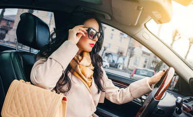 Fashion girl with glasses sits behind the wheel of a premium car and looks the way