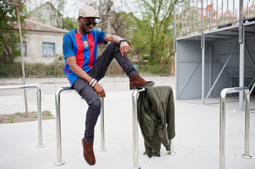 Stylish african american boy wear at cap, football t-shirt and sunglasses posed on steel railings. Black sports man portrait.