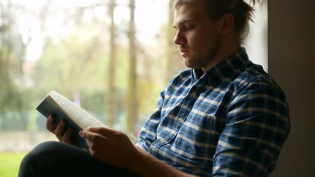 Handsome man sitting next to the window and reading book
