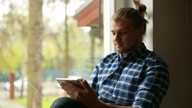 Handsome man with mud bun sitting next to the window and using tablet
