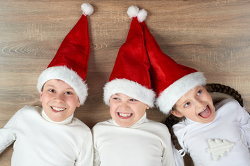 three kids in Santa hats lying on wooden background, having fun and happy emotions, winter holiday concept