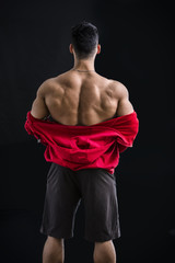 Obraz na płótnie Canvas Muscular bodybuilder undressing, removing red hoodie sweater on naked muscle back on dark background