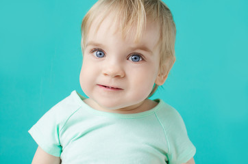 Naklejka premium A little girl (boy) with blue eyes in a light green T-shirt is looking at the camera in the color of the sky background. Demanding expression face. An unusual emotion.
