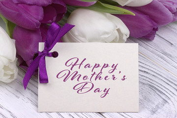 Purple and white tulips with white paper on a white wooden background and card lettering happy Mother's day.