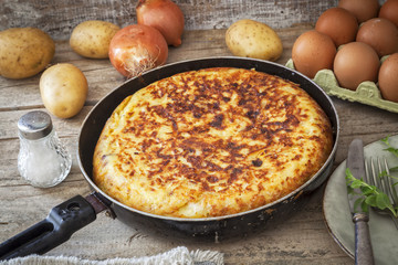 Homemade Spanish potato omelet in a frying pan on a wooden table with ingredients.