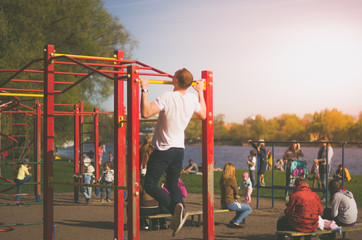 A man is practicing on a bar in a public park. A man pulls himself up on a bar in a street playground