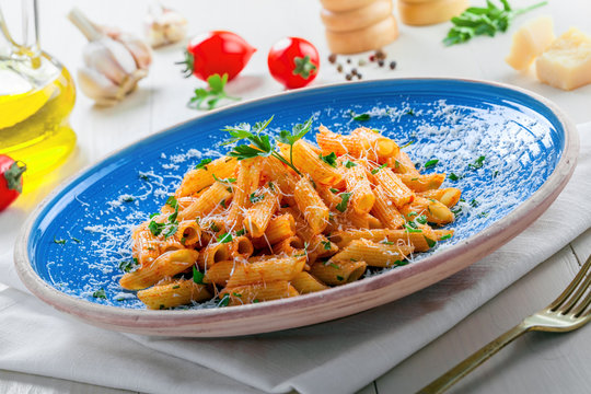 Traditional penne pasta with hot arrabiata tomato sauce and parsley on a white wooden table. Traditional Italian healthy food on a blue rustic plate. Close-up.