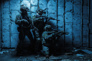 Squad of Army Rangers with rifle and machine gun moving along the concrete wall of ruined destroyed building on mission. They are ready to start firing if enemy appear. Outdoor location shot, darkness