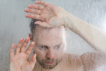stressed man standing under flowing water and puts his hand on the glass in shower cabin behind transparent misted glass door in the bathroom