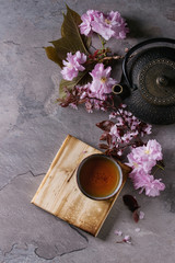 Obraz na płótnie Canvas Black iron teapot and traditional ceramic cup of tea on wooden serving board with blossom pink flowers cherry branch over gray texture background. Top view with space, Asian style.