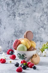 Variety of colorful french sweet dessert macaron macaroons with different fillings served in vintage tea cup with spring flowers and fresh berries over gray texture background.