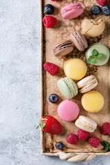 Variety of colorful french sweet dessert macaron macaroons with different fillings served on terracotta tray with fresh berries over gray texture background. Top view with space