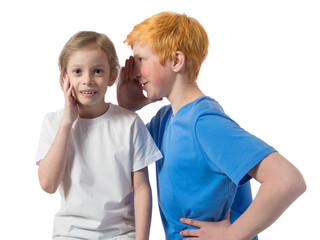 Fototapeta premium Little boy in a blue t-shirt whispers in the ear to a surprised girl, the girl holds his hand behind his cheek against a white background
