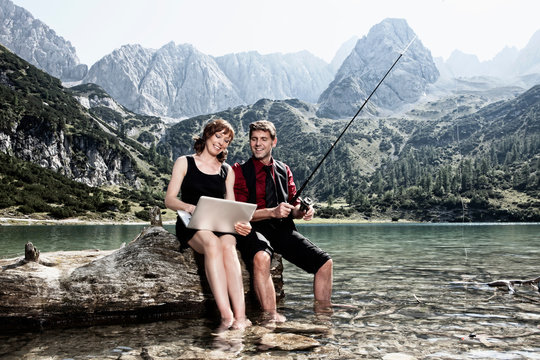 Couple fishing and using laptop by lake