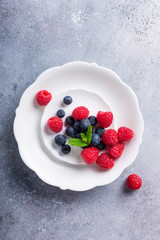 Freshly picked blueberries and raspberries on white plate on gray stone background. Concept for healthy eating and nutrition with copy space. Top view.