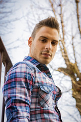 An attractive young man sitting on ground outdoor in public park against metal fence, wearing jeans and shirt