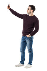 Side view of handsome casual man with sunglasses taking selfie with cell phone. Full body length portrait isolated over white studio background.