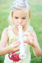 Blond child girl playing flute outdoors in the garden with green grass lawn background