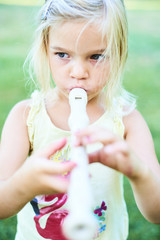 Blond child girl playing flute outdoors in the garden with green grass lawn background
