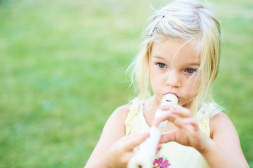 Blond child girl playing flute outdoors in the garden with green grass lawn background