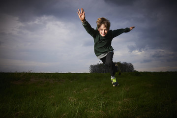 Child blond boy jumping in a spring green meadow against a storm's cloudy sky at sunset
