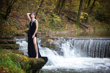 Portrait of a beautiful brunette in a long black dress on the river bank
