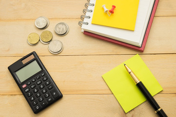 top view. pen putting on yellow post-it note and have black calculator, coin, pin and more post-it note putting on notebook. wooden are background. this image for business, 