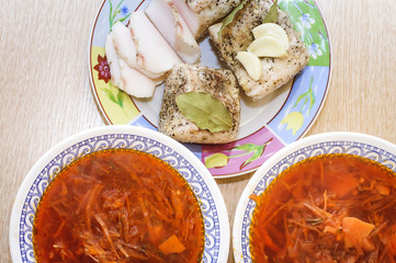 Salted lard with pepper, bay leaf and garlic lies on a plate, next to it there are two plates with red borsch.. Close-up, view from above.