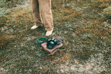 Man standing next to leather bag with gloves and camera lying in field. High angle view.