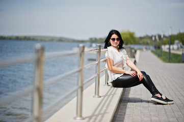 Portrait of  brunette girl on women's leather pants and white blouse, sunglasses, against iron railings at beach.