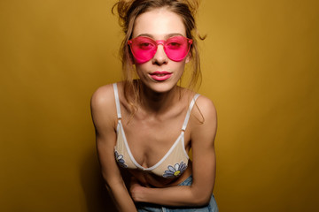 The girl with collected hair and the top of the swimsuit in the pink glasses is on a yellow background leaning forward and looking at the camera