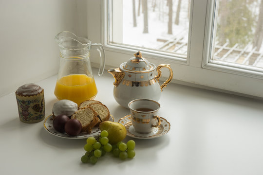 Still life with  Easter cakes, eggs, teapot ,  cup of tea juice, grapes and pear  on a sill and trees covered with snow seen through the open window