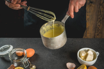 Traditional basic sauces. French cuisine. Person in the frame is preparing Hollandaise sauce. In a metal saucepan, with ingredients - eggs, butter, lemons. On black stone table. Female hands.
