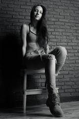 Black-and-white photo of a girl in top and jeans sitting on a stool against a brick wall, view from below. Vertical photo