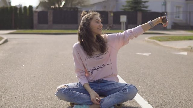 Cute hipster girl makihg selfie with skateboard outdoors in sunset light.