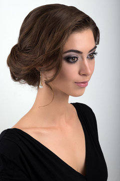 Close-up portrait of brunette with evening makeup and collected hair in a black dress standing half-turn on a light background