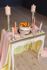 Wedding cake with orange berries, candles and two glasses stand on a white table decorated with a pink cloth
